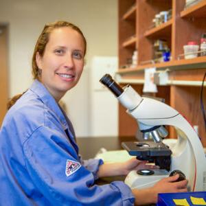 泰莎·所罗门-莱恩(Tessa Solomon-Lane)坐在实验室的显微镜前，对着镜头微笑. 她长长的深金色头发向后梳着，穿着一件蓝色的实验服.